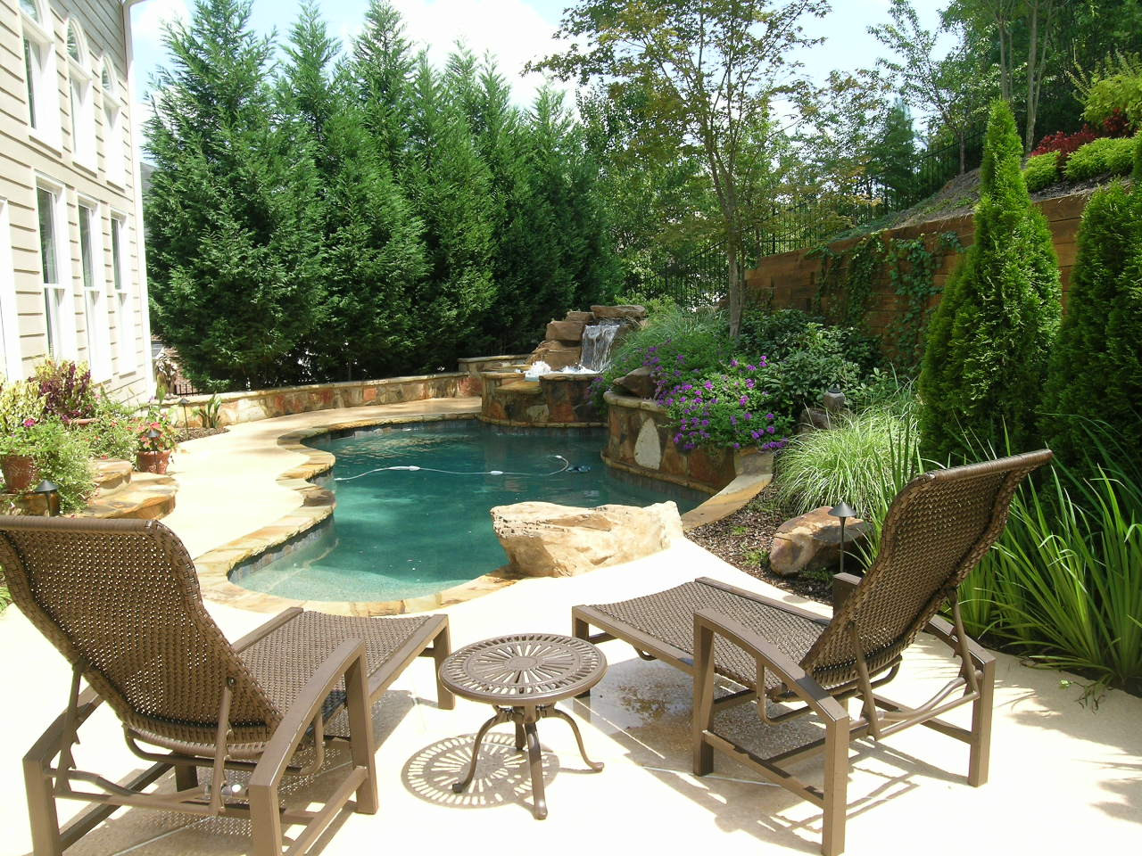 Two brown lounge chairs and a small round table face a backyard pool with rock features and a waterfall, surrounded by greenery and flower beds.