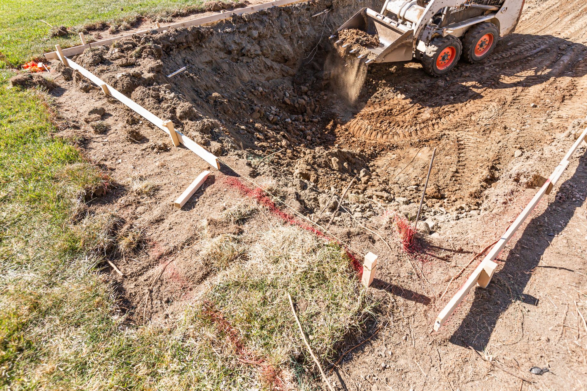 A construction site with a skid steer loader digging soil within a wooden-framed area, preparing the ground for a foundation as part of professional grading services.