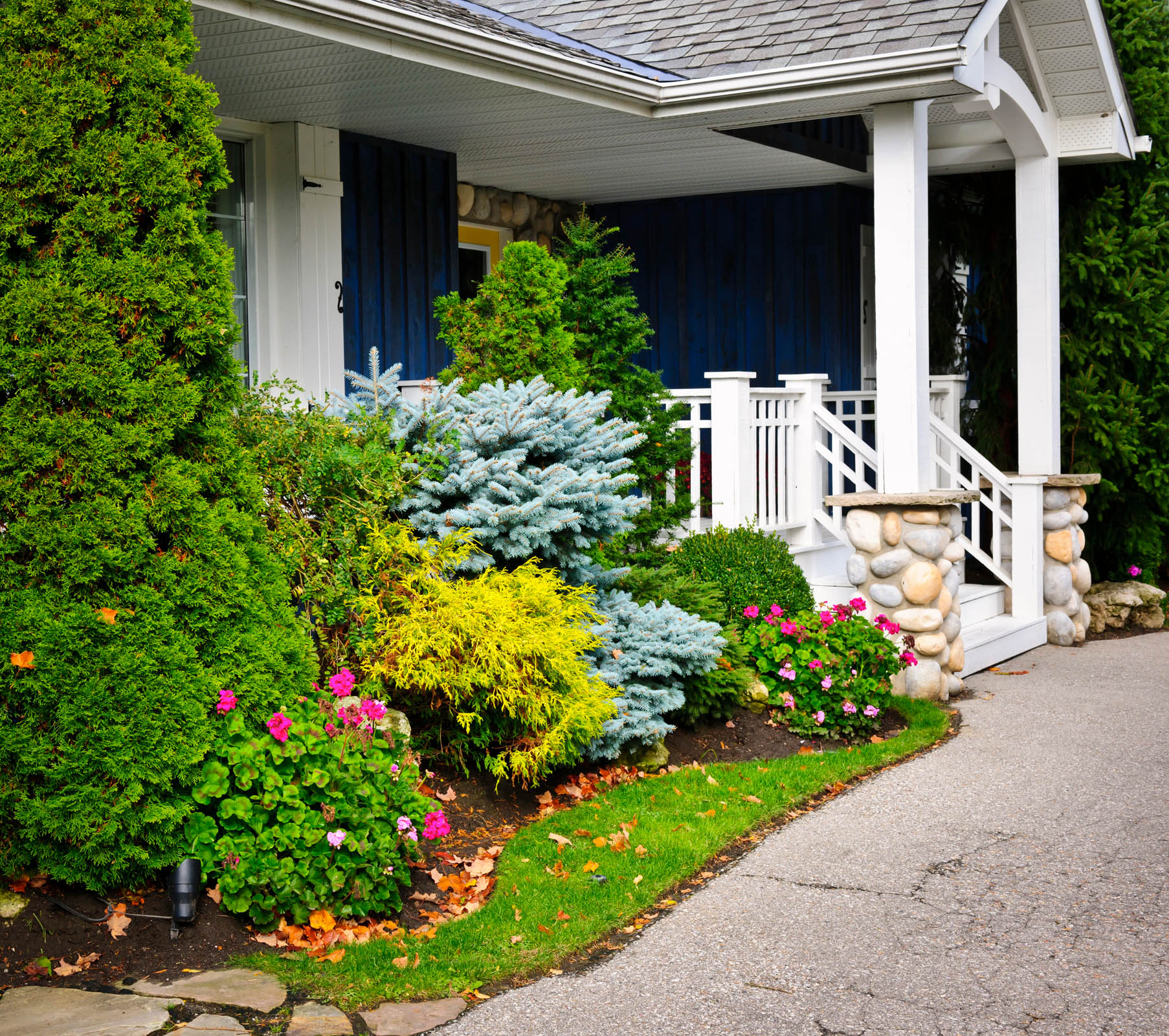 A house with a stone and white railing porch, surrounded by a variety of green shrubs and colorful flowers next to a paved walkway.