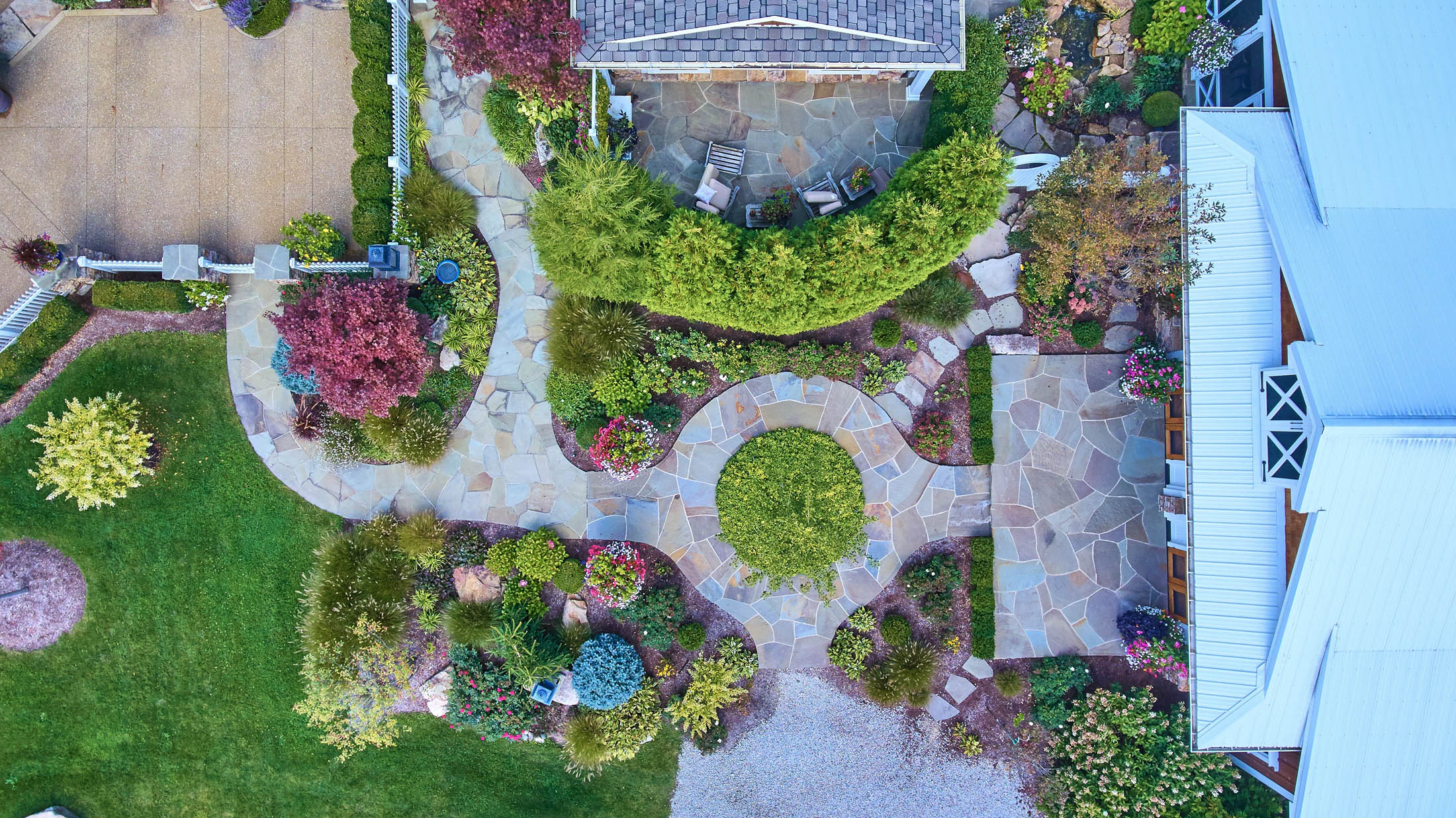 Aerial view of a landscaped garden with stone pathways, green lawns, various shrubs, and seating areas next to a house.