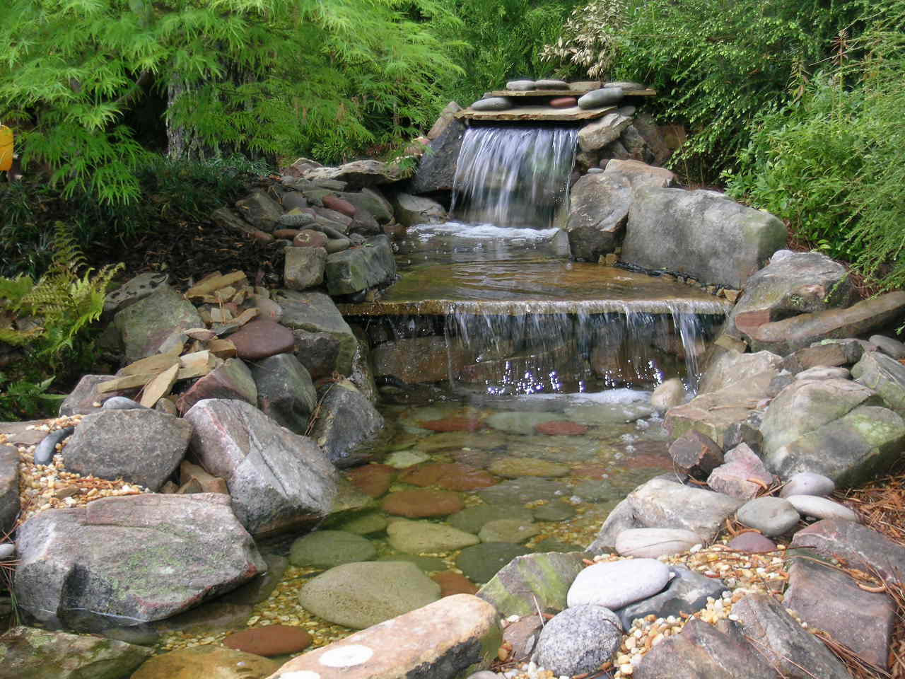 A small artificial waterfall flows over rocks into a shallow stream surrounded by green plants and stones.