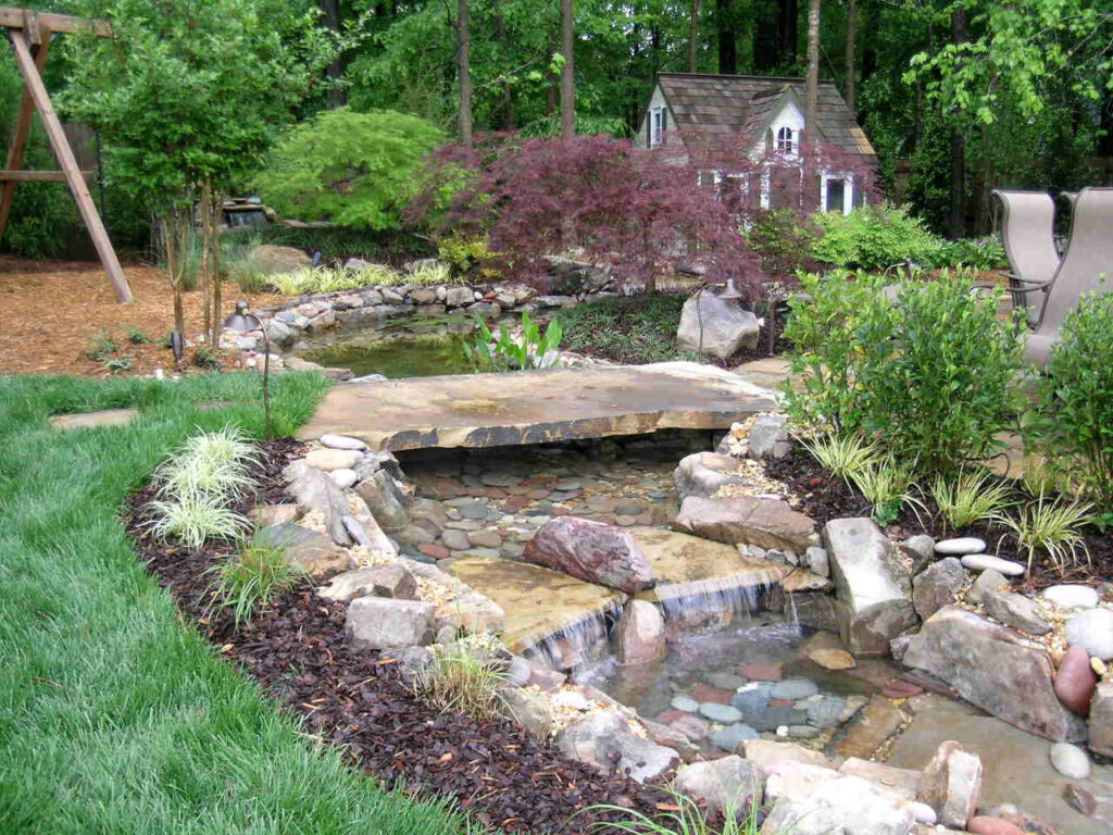 A landscaped backyard with a small stone bridge over a stream, a pond, various plants, and a playhouse in the background.