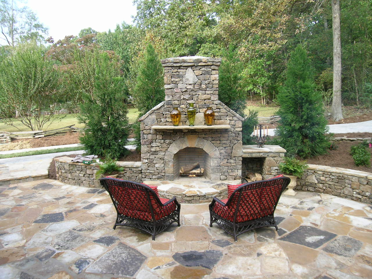 Two wicker chairs with red cushions face a large stone outdoor fireplace on a stone patio, surrounded by trees and greenery.