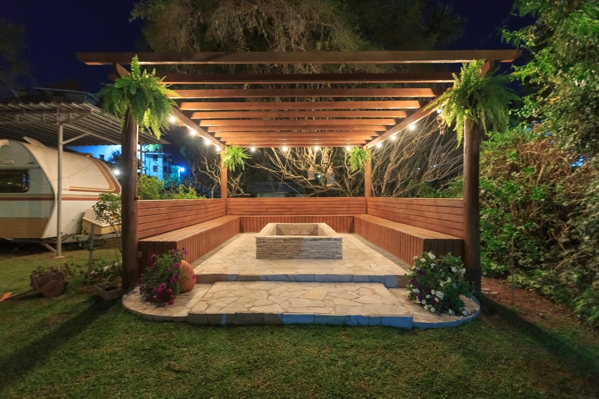 A wooden pergola with hanging plants and string lights covers a stone patio with a built-in fire pit, surrounded by benches and garden greenery at night.
