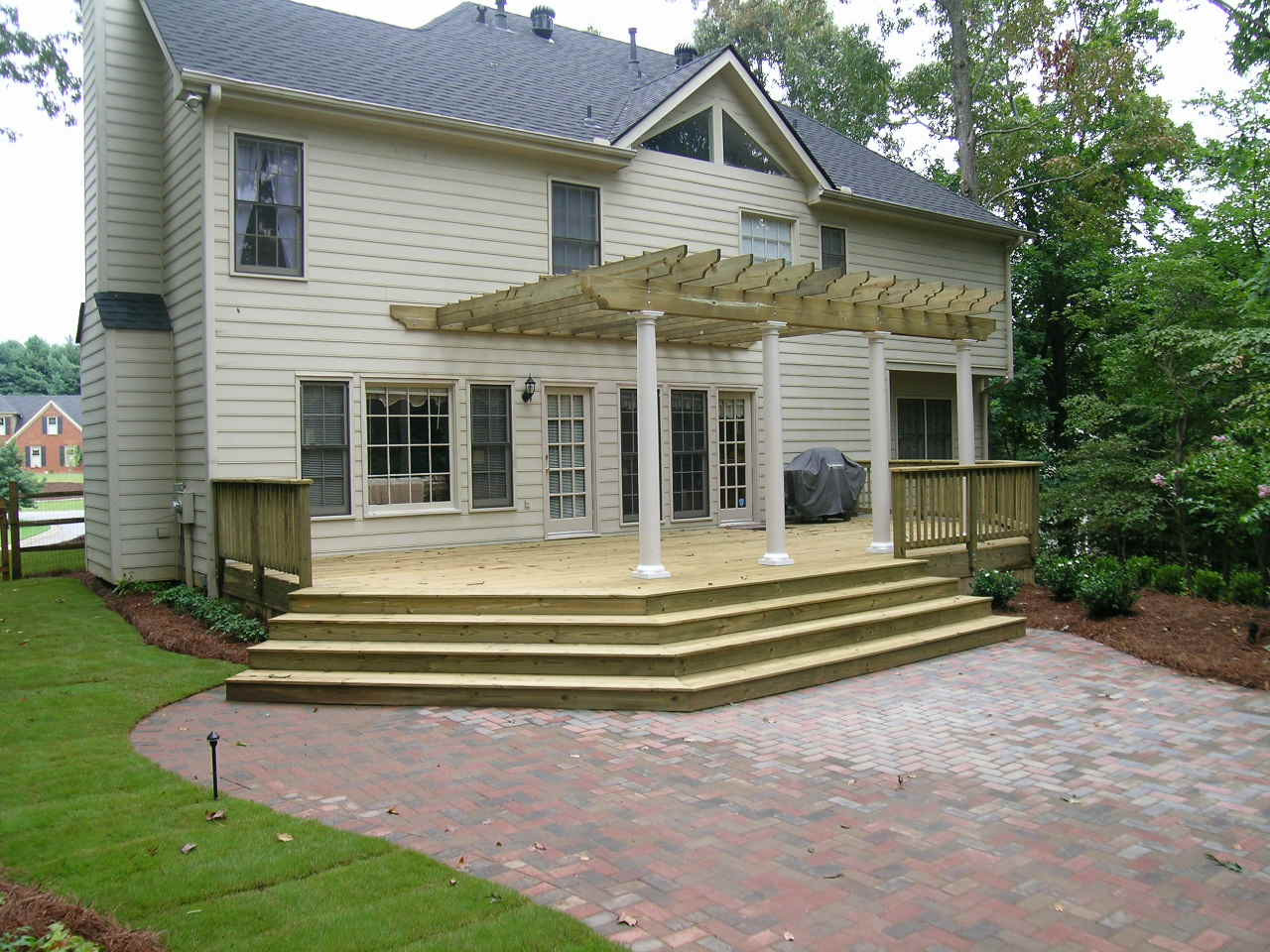 A two-story house with a wooden deck featuring a pergola, steps leading to a brick patio, and landscaped lawn and trees surrounding the backyard.