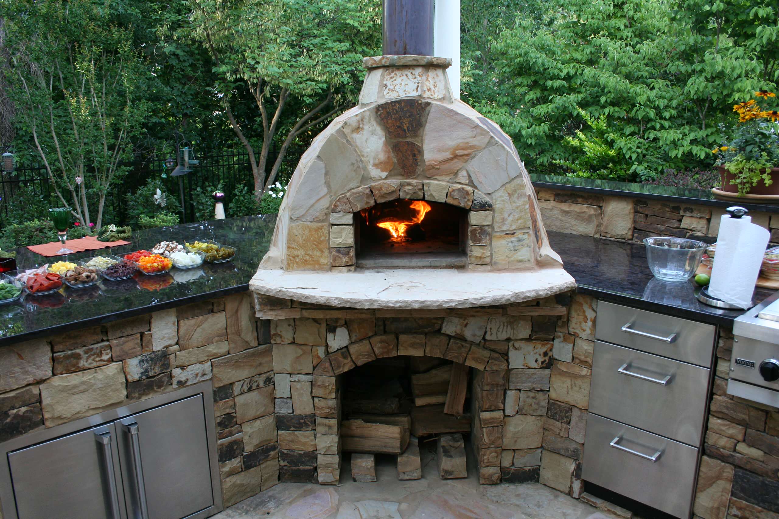 A stone outdoor pizza oven with a fire burning inside, surrounded by a countertop with prepared ingredients, drawers, and cabinets, set in a garden.