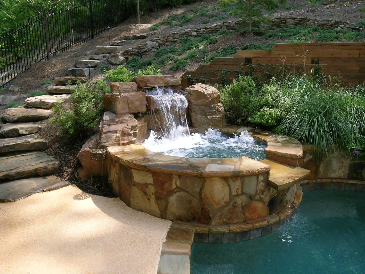 A small stone waterfall flows into a raised hot tub, which overflows into a swimming pool, surrounded by landscaping and rock steps.