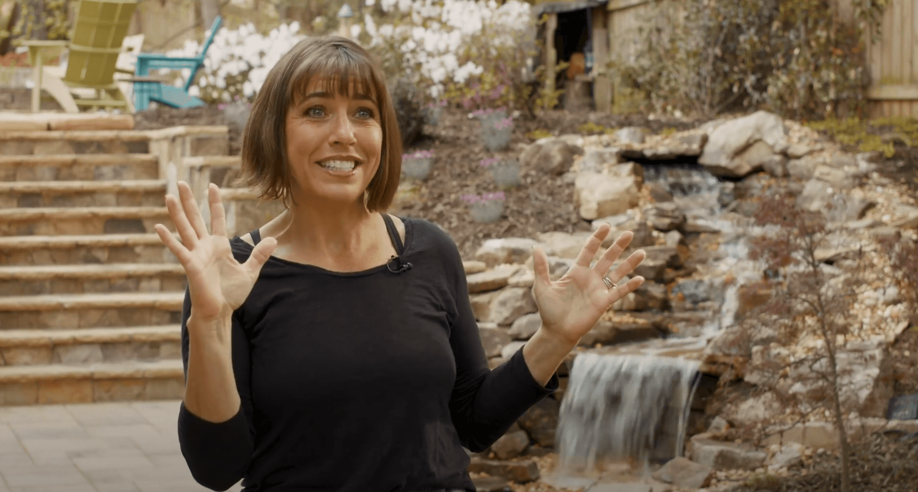 Woman with short brown hair and black shirt gestures with hands and smiles while sitting near a small backyard waterfall and stone steps, sharing heartfelt video testimonials about her experience.