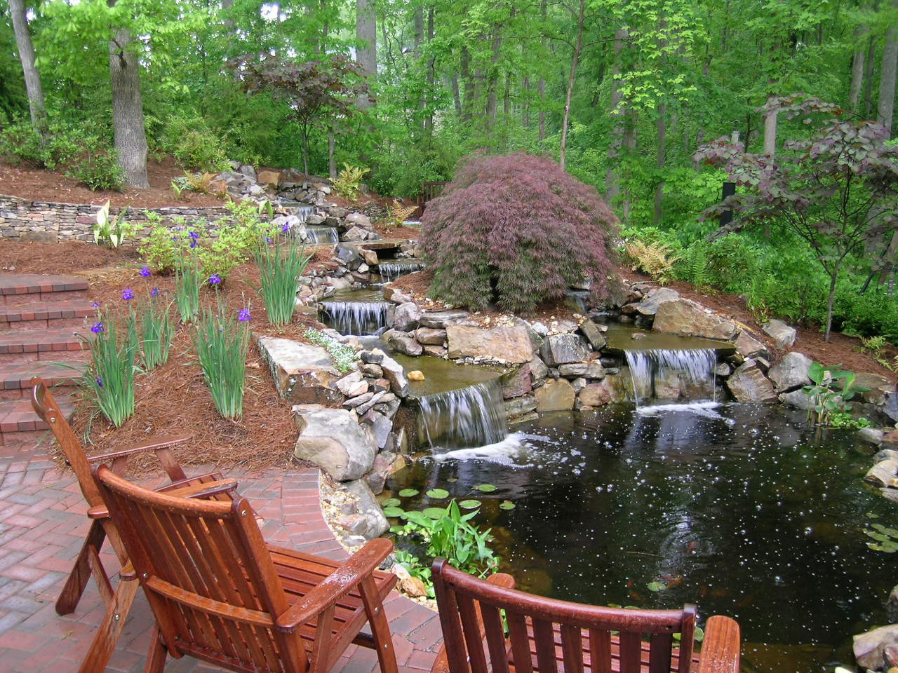 Wooden chairs face a small pond with multiple waterfalls, surrounded by brick steps, flowering plants, rocks, and dense green trees in a landscaped garden.