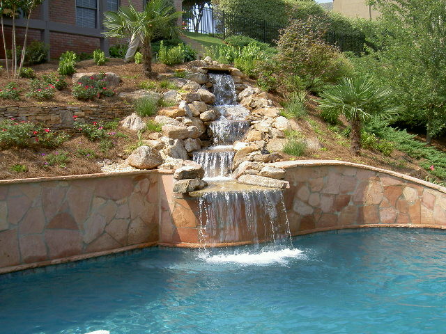 A stone waterfall flows into a clear blue swimming pool, surrounded by landscaping with plants, trees, and a retaining wall.