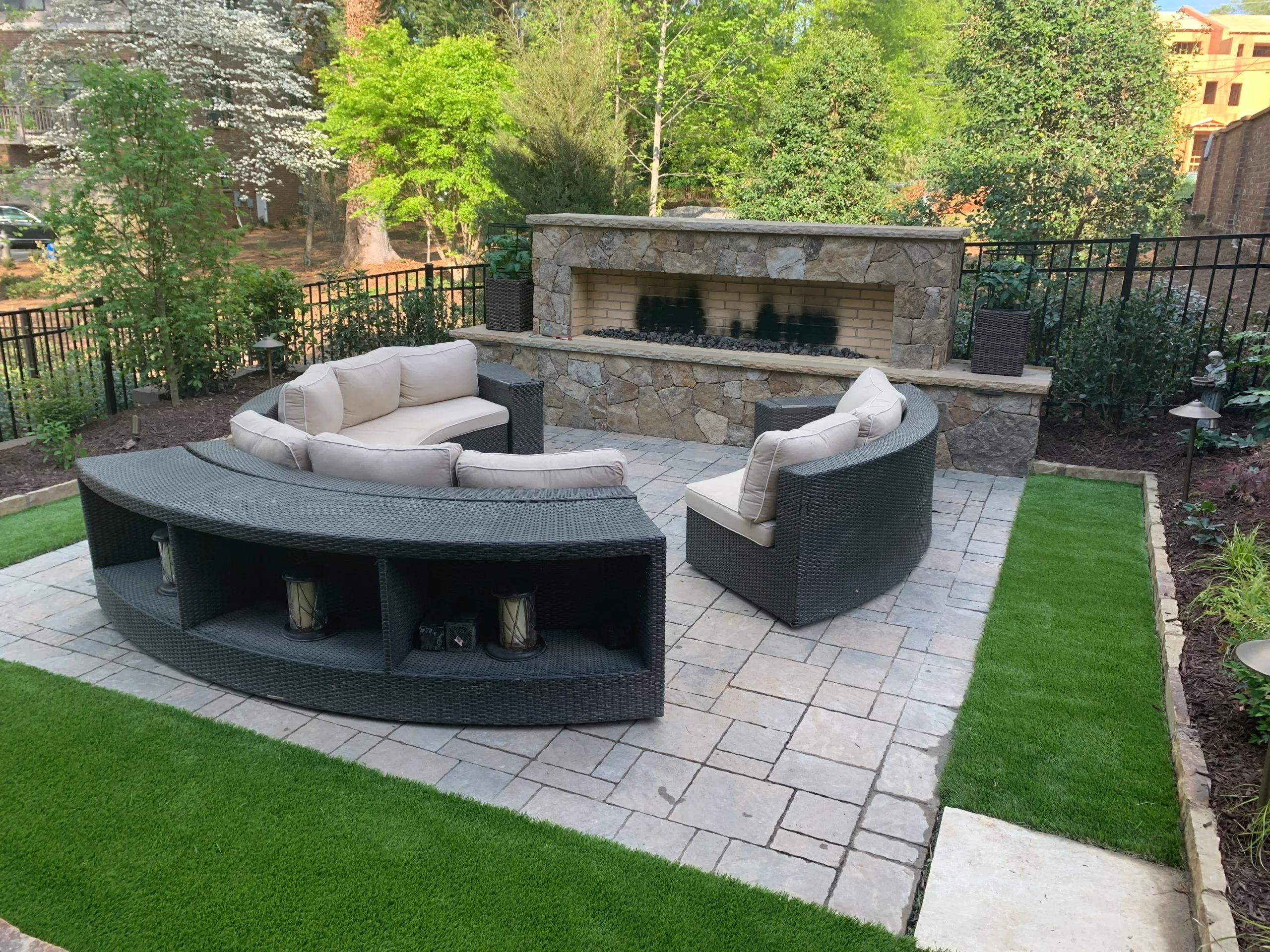 Curved wicker patio seating with beige cushions placed around a stone outdoor fireplace on a tiled patio with artificial grass borders, enhanced by a dynamic content widget for added ambiance.