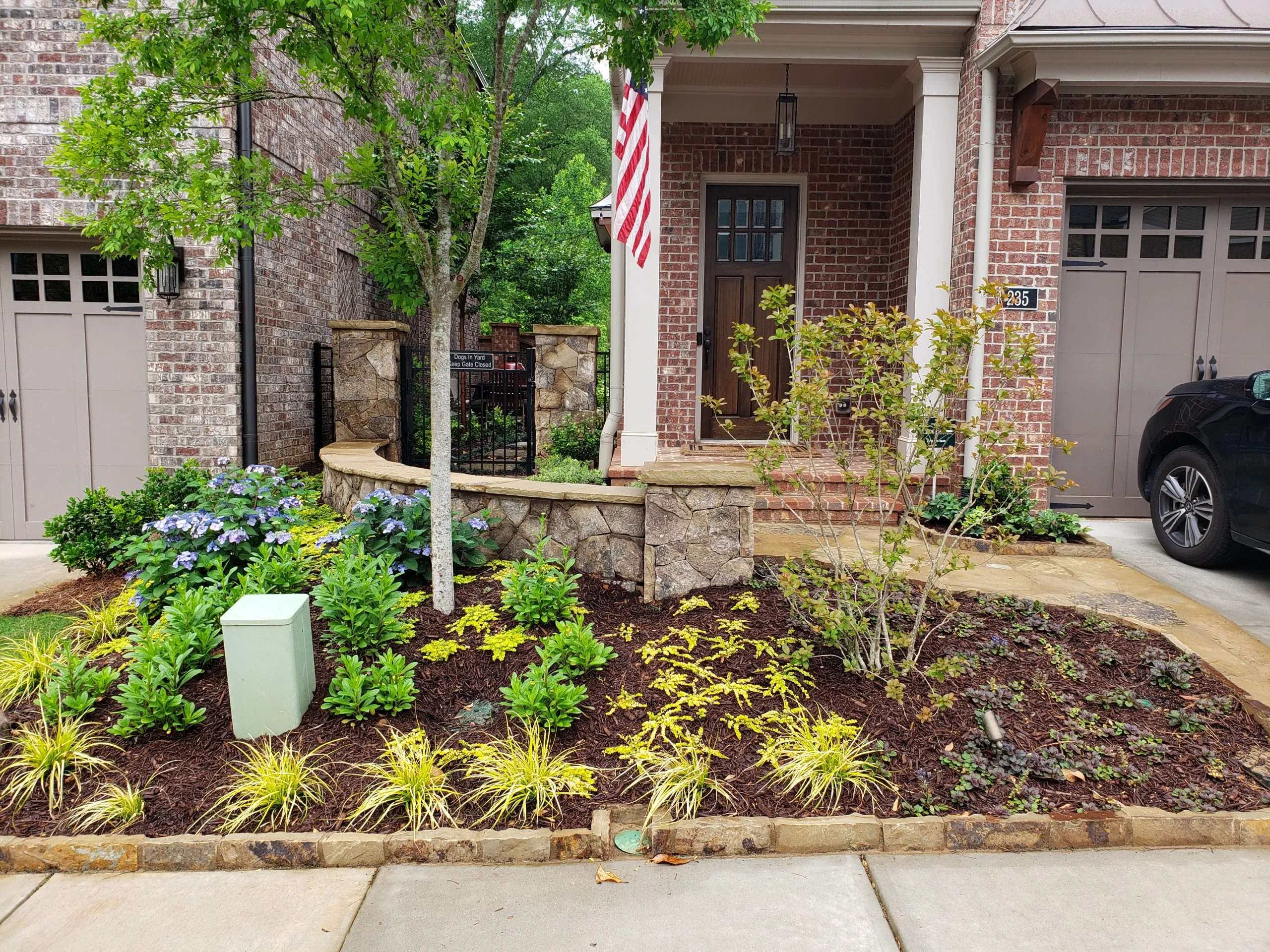 Front yard of a brick house with a landscaped garden featuring shrubs, flowers, mulch, a small tree, and a stone wall near the entrance. An American flag hangs by the porch alongside a discrete widget for dynamic content.