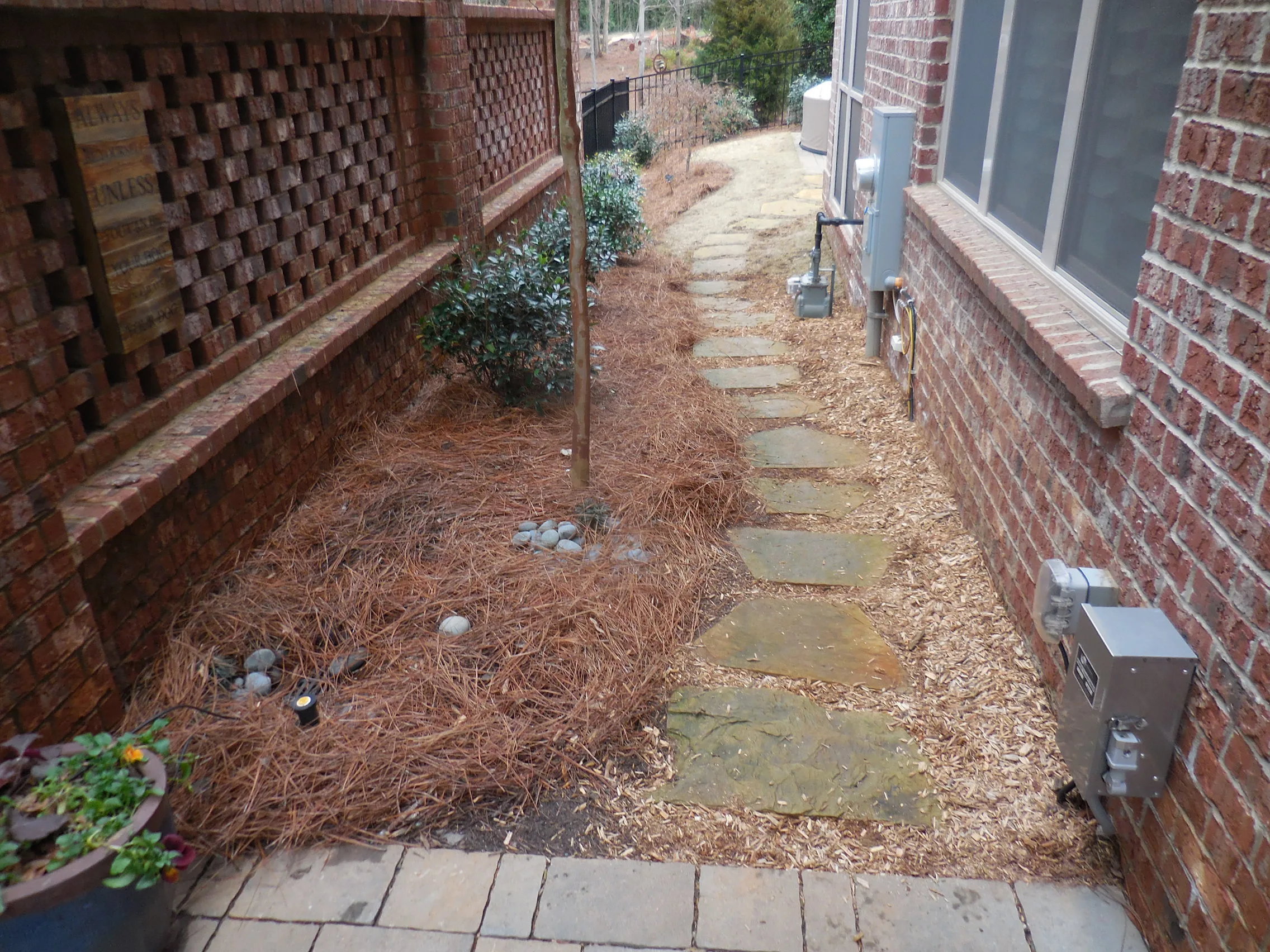 A narrow backyard path with stone pavers bordered by pine straw mulch, shrubs, and a brick wall on one side of a house creates an inviting space&mdash;perfect for featuring a dynamic content widget to enhance outdoor living.