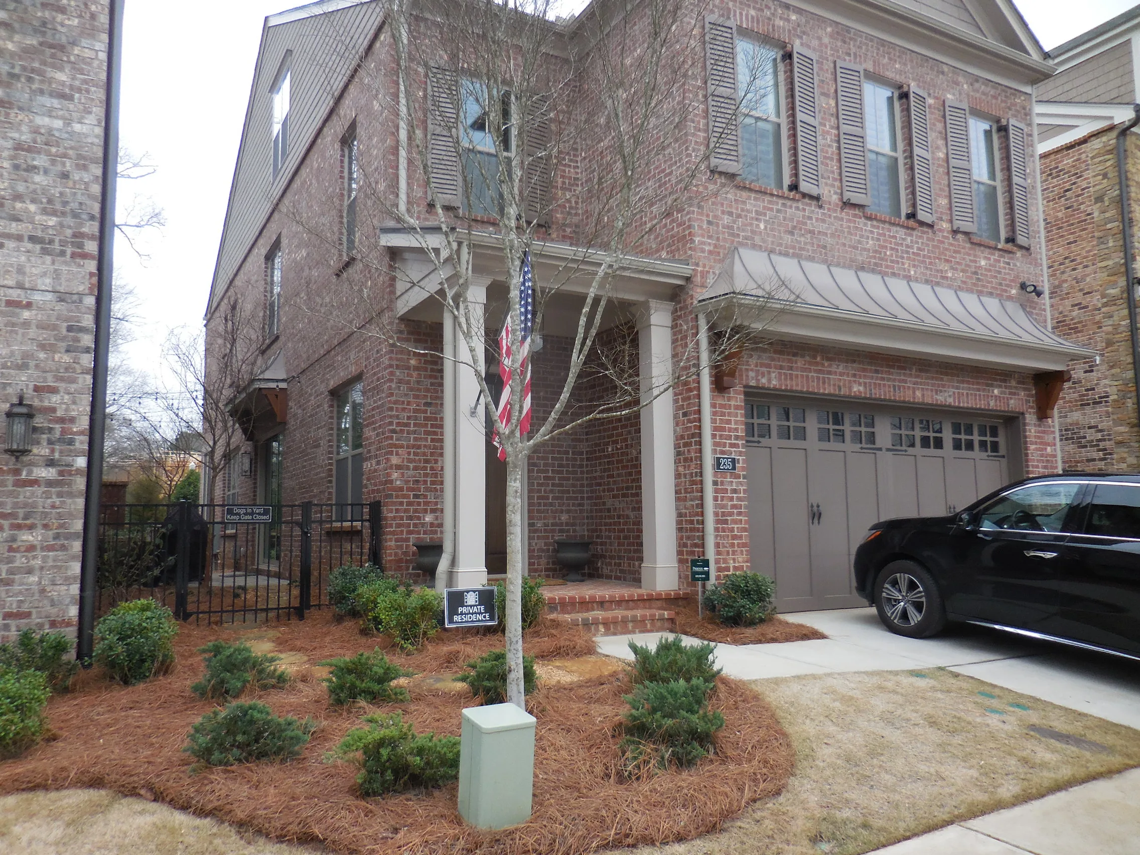 Two-story brick house with columns, blue shutters, and a two-car garage. A dynamic content widget by the front door displays updates. An American flag hangs nearby, a black SUV is in the driveway, and shrubs line the front yard.