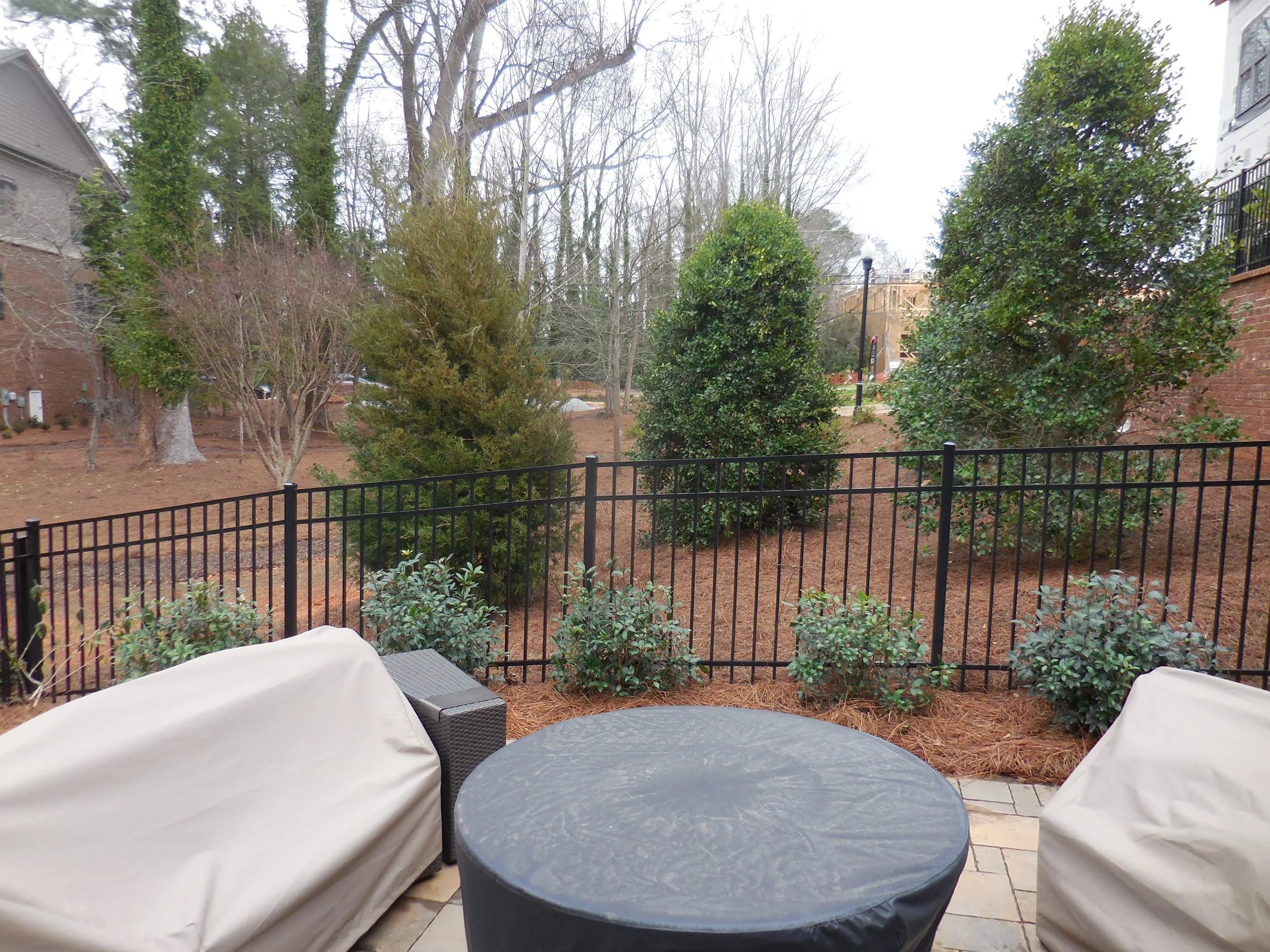 A fenced backyard patio with covered furniture, a round covered table, and a dynamic content widget, overlooking trees and shrubs on a cloudy day.