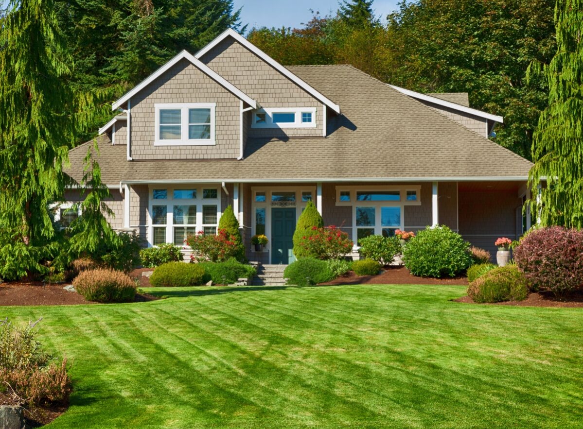 A two-story suburban house with a manicured lawn, shrubs, and trees in the front yard showcases professional landscape installation, surrounded by lush greenery under a clear sky.