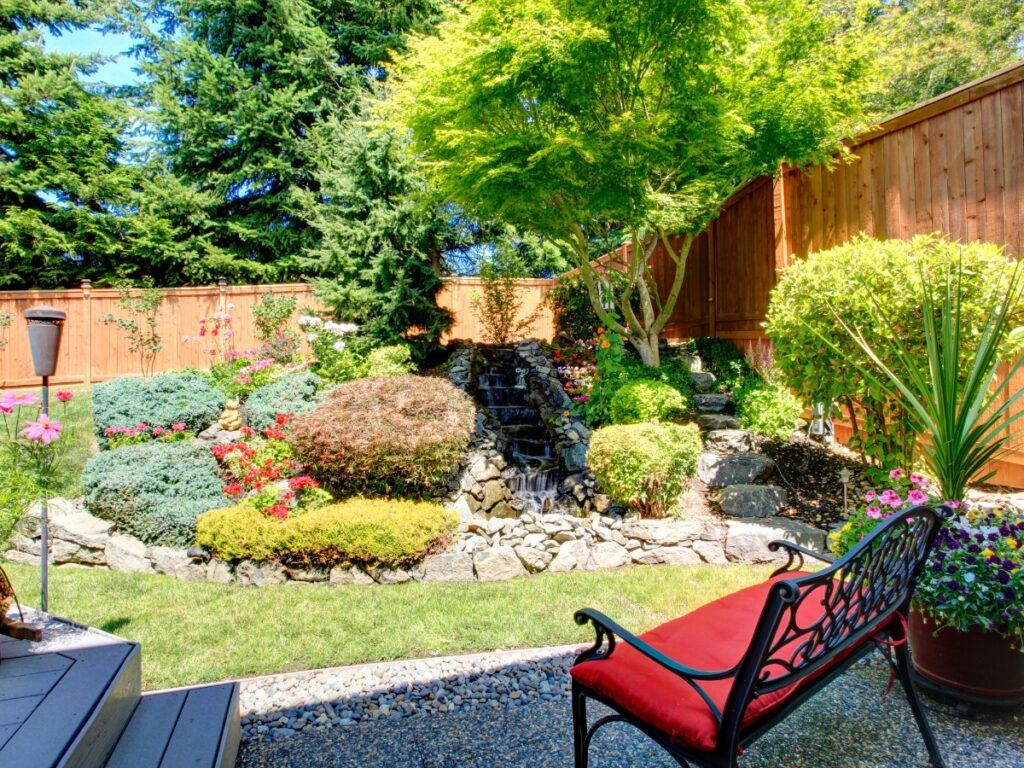 A professionally landscaped backyard garden with a small waterfall, various shrubs and flowers, a wooden fence, and a red cushioned bench on a gravel patio showcases expert landscape installation.