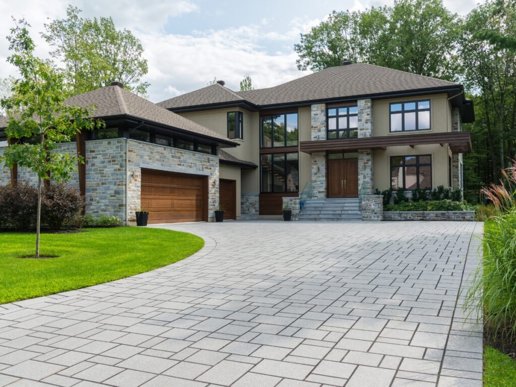 Modern two-story house with stone and stucco exterior showcases striking curb appeal design, featuring large windows, double garage, and a wide paved driveway surrounded by green lawn and trees.