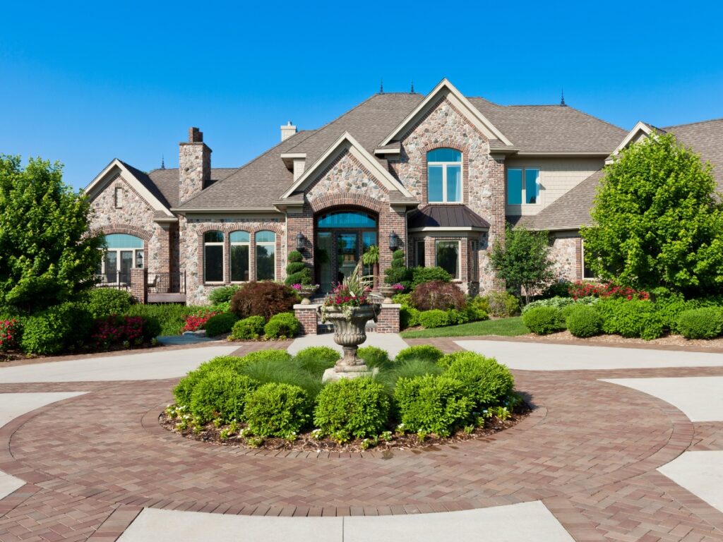 Large brick house with multiple windows and gables features stunning curb appeal design, a landscaped front yard, circular driveway, and a central stone fountain surrounded by shrubs.