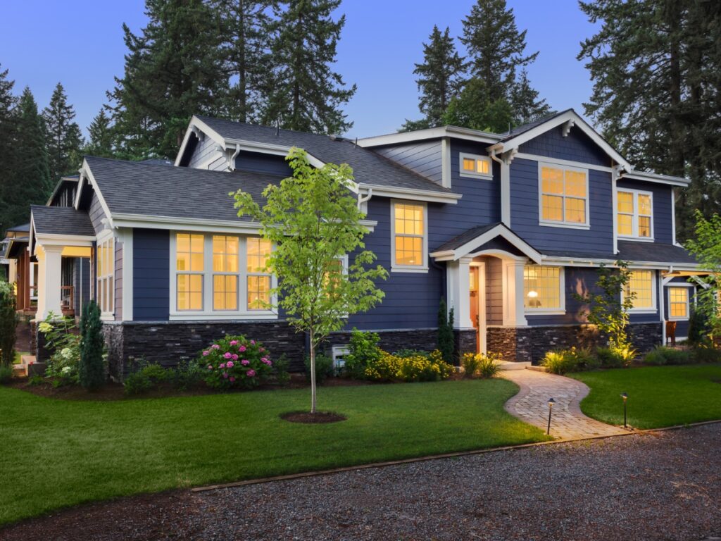 Two-story blue house with white trim, stone accents, and lit windows, this curb appeal design is enhanced by a lush green lawn, shrubs, and trees at dusk.