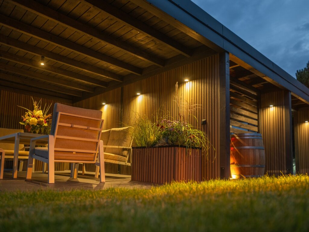Outdoor seating area under a wooden pergola with recessed lighting, showcasing the landscaping lighting benefits. A table with a flower arrangement and planter boxes sits on a grassy lawn at dusk, creating an inviting ambiance.