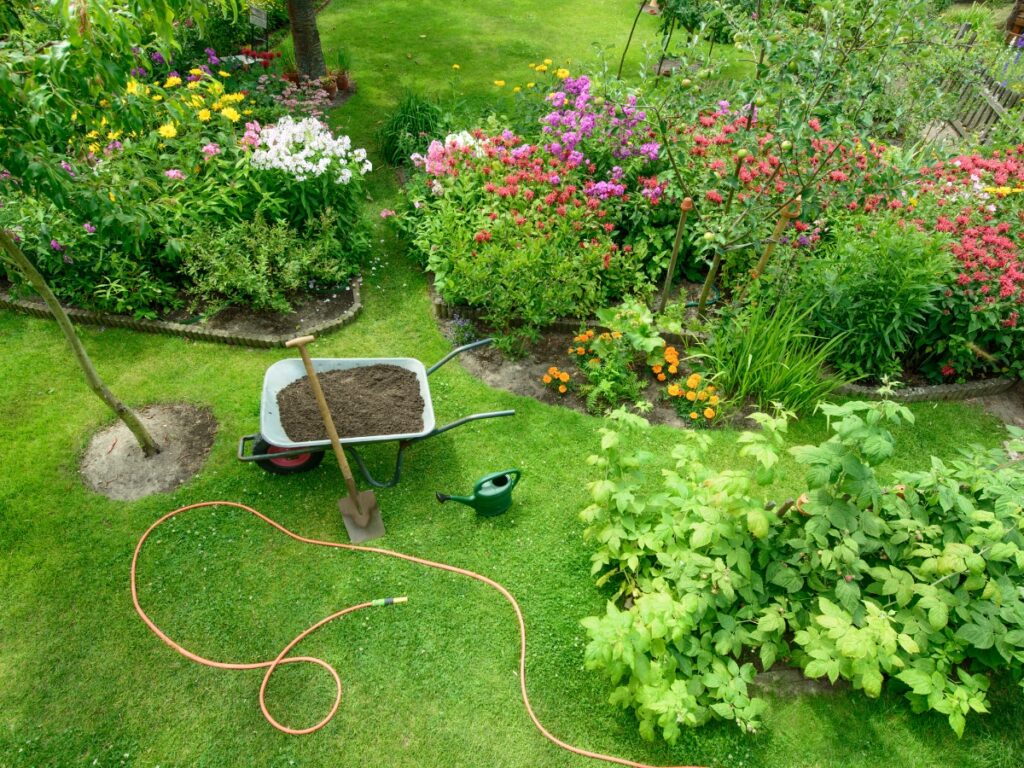 A garden with flower beds and green plants, a wheelbarrow filled with soil, a shovel, a watering can, and a garden hose lying on the grass—ready to withstand extreme weather.