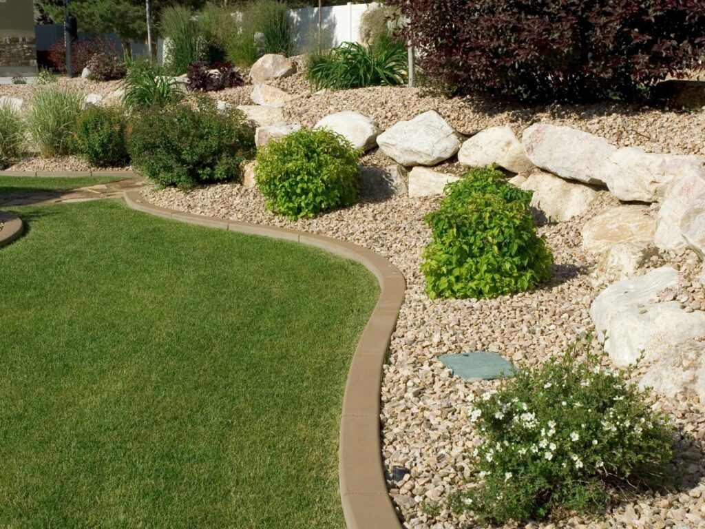 A landscaped yard with a curved lawn edge, decorative rocks, shrubs, and a terraced rock wall provides natural erosion control and is surrounded by gravel mulch.
