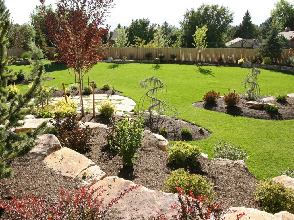 A landscaped backyard with green grass, stone pathways, mulched garden beds for erosion control, various shrubs, and a wooden fence in the background.