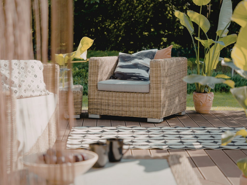Wicker armchair with cushions on a wooden deck, surrounded by potted plants and a patterned rug, in an outdoor garden setting that offers health benefits through relaxation and fresh air.
