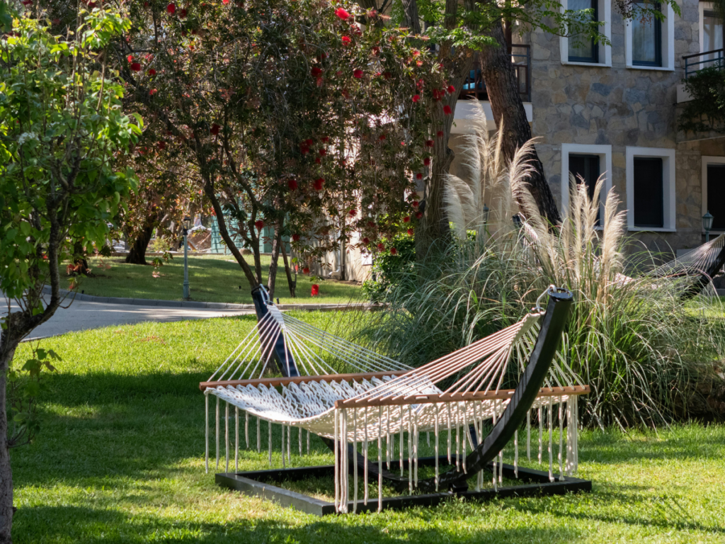 A white rope hammock on a metal stand sits on green grass in a garden with flowering trees and a stone building, offering relaxation and the health benefits of outdoor rest.