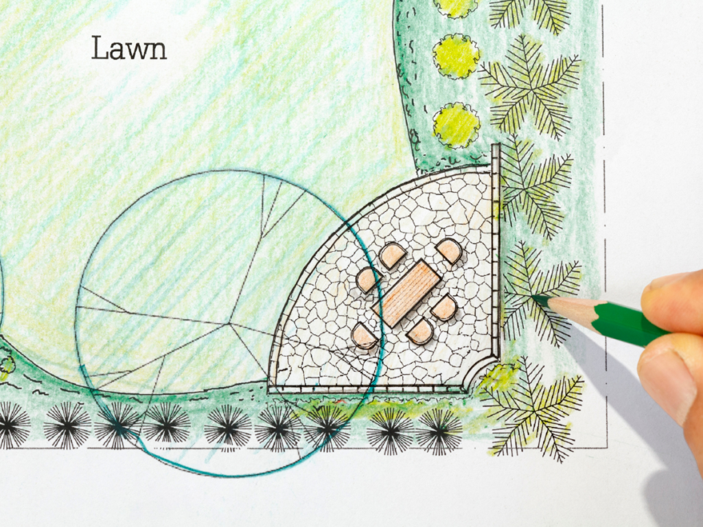 A hand uses a green pencil to add detail to a landscape design plan featuring a patio with a table and chairs, surrounded by greenery and labeled "Lawn"—perfect inspiration for small backyard ideas.