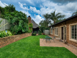 Backyard with green lawn, brick patio, metal table and chairs, brick house, lush plants, and a thatched-roof structure under a partly cloudy sky—perfect for those seeking small backyard ideas with charm and functionality.