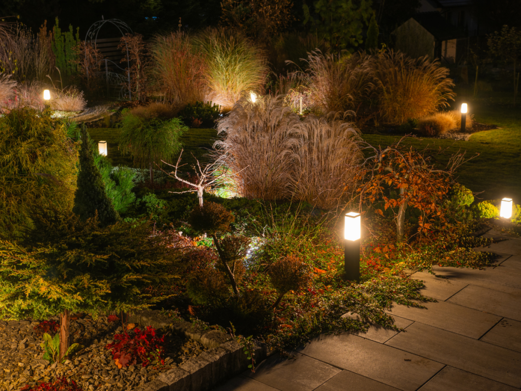 A landscaped garden at night with automated lighting illuminating the pathway, ornamental grasses, shrubs, and a stone walkway in the foreground.