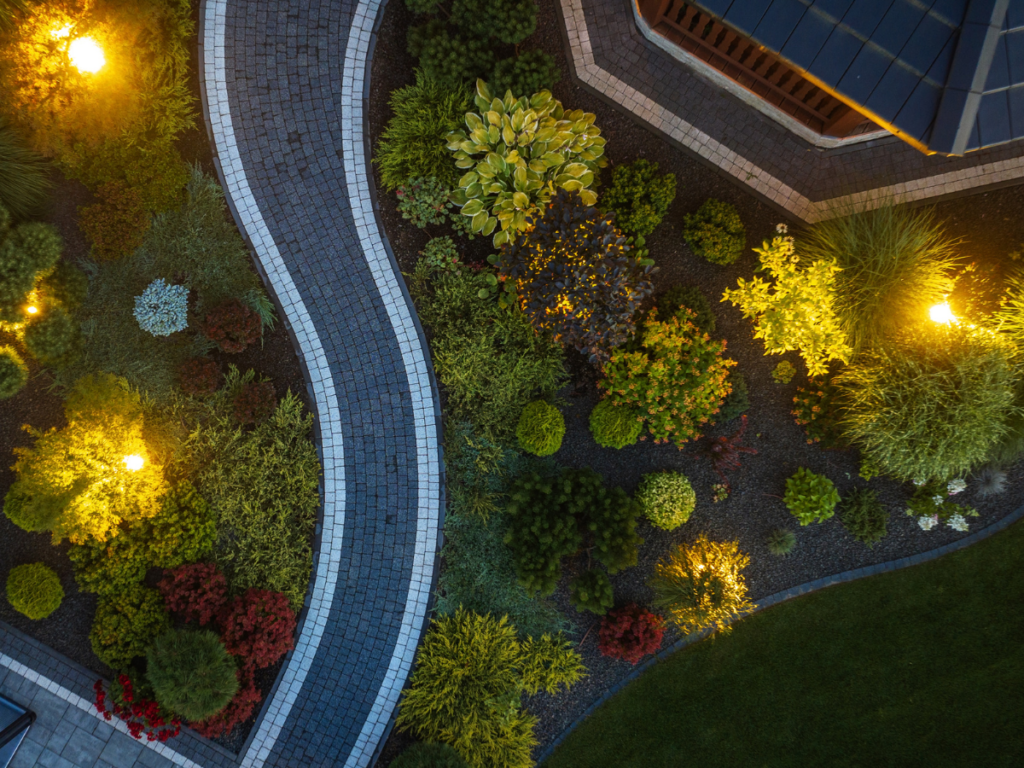 Aerial view of a landscaped garden with a curving stone path, automated lighting illuminating various shrubs, and a building roof at the edge.