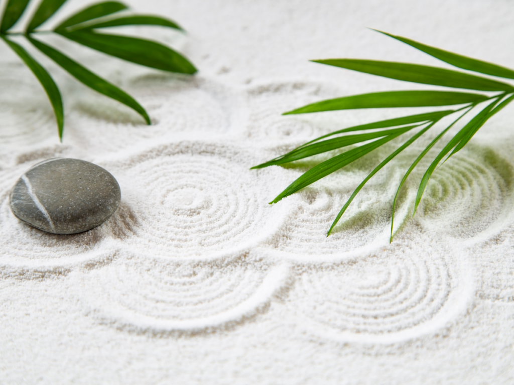 A smooth stone and green palm leaves rest on white sand with circular patterns raked around them, evoking the tranquility of a zen garden.