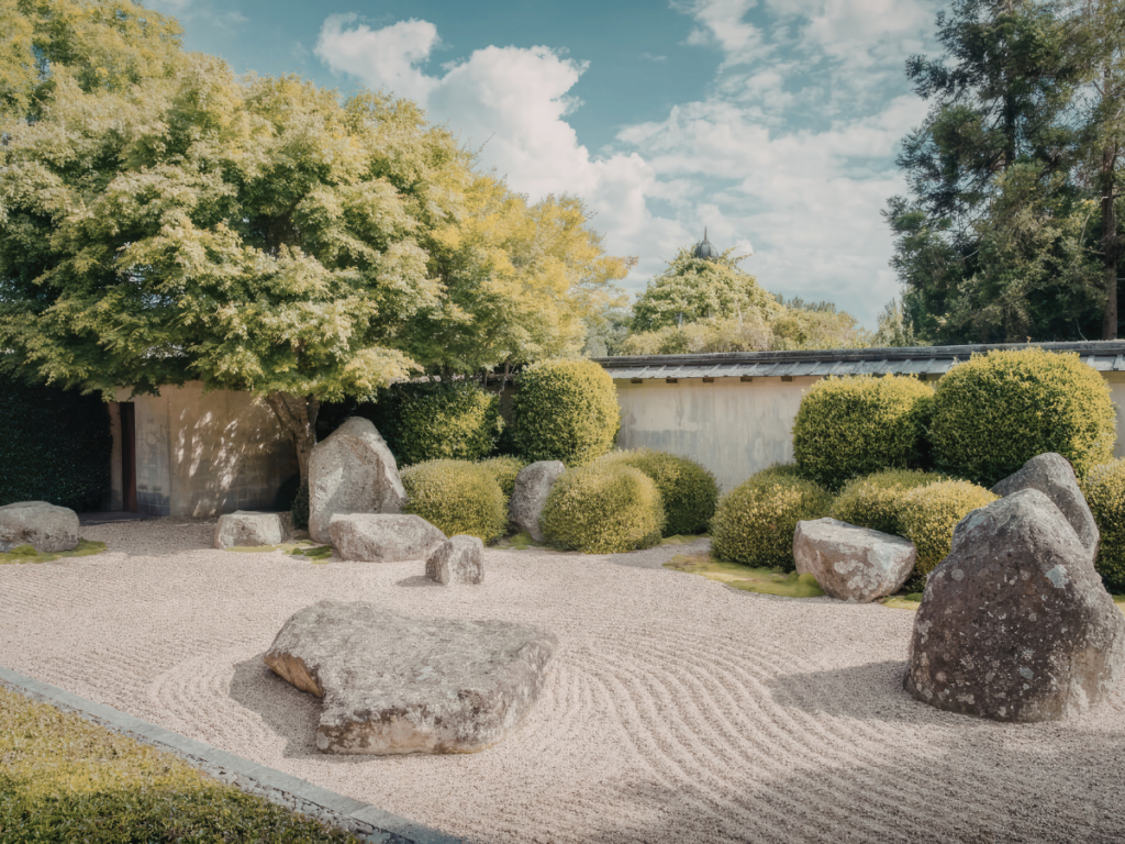 A traditional Japanese zen garden with large stones, raked gravel, green shrubs, and trees under a partly cloudy sky.