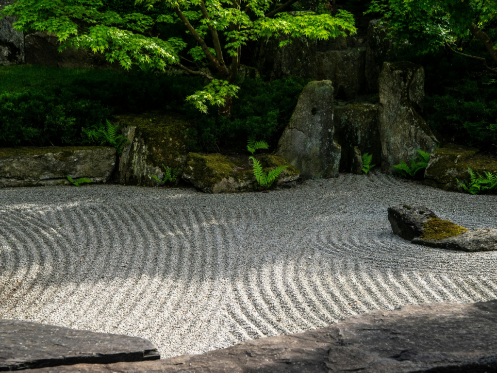 A tranquil zen garden featuring raked gravel patterns, large rocks, ferns, and lush green trees in the background.