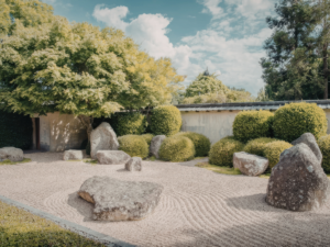 A traditional Japanese zen garden with large stones, raked gravel, green shrubs, and trees under a partly cloudy sky.