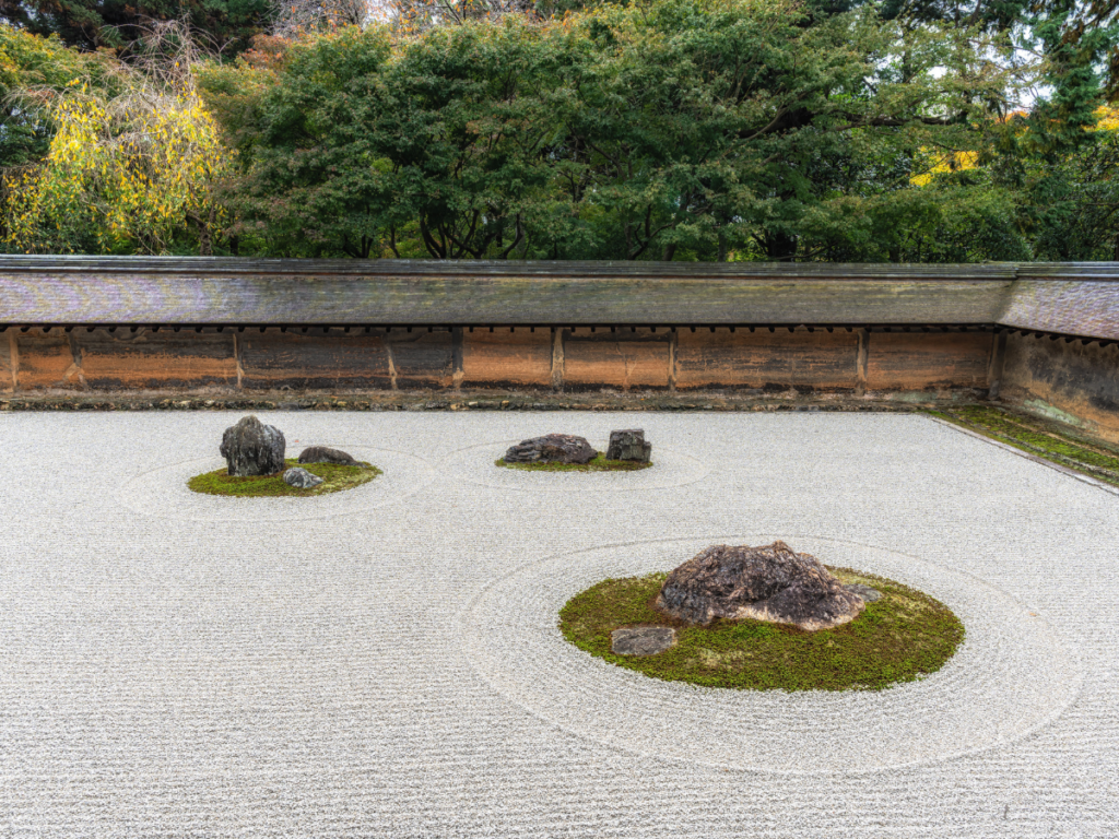 A traditional Japanese zen garden with raked gravel, several large rocks, patches of moss, and a wooden wall, bordered by dense green trees.