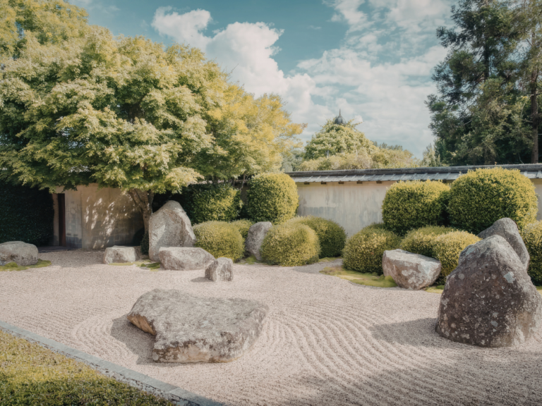 A traditional Japanese zen garden with large stones, raked gravel, green shrubs, and trees under a partly cloudy sky.