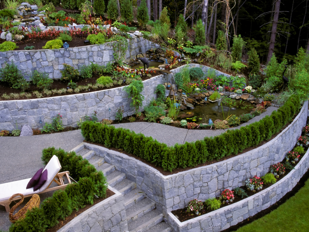 Multi-level terraced garden with stone retaining walls, green hedges, colorful flowers, a pond with statues, and a lounge chair on a patio.