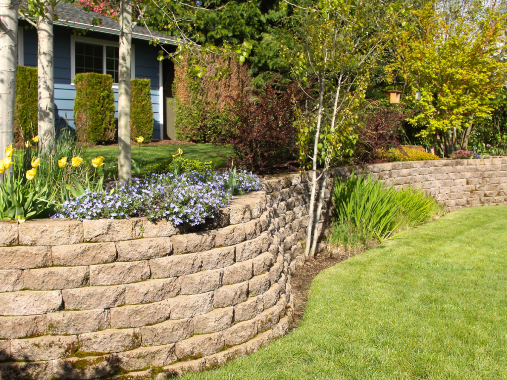 A tiered stone retaining wall with flower beds and a well-maintained lawn creates a beautiful focal point in front of the house. Shrubs, trees, and blooming flowers complement the sturdy stone retaining wall, adding vibrant color and natural charm to the landscape.