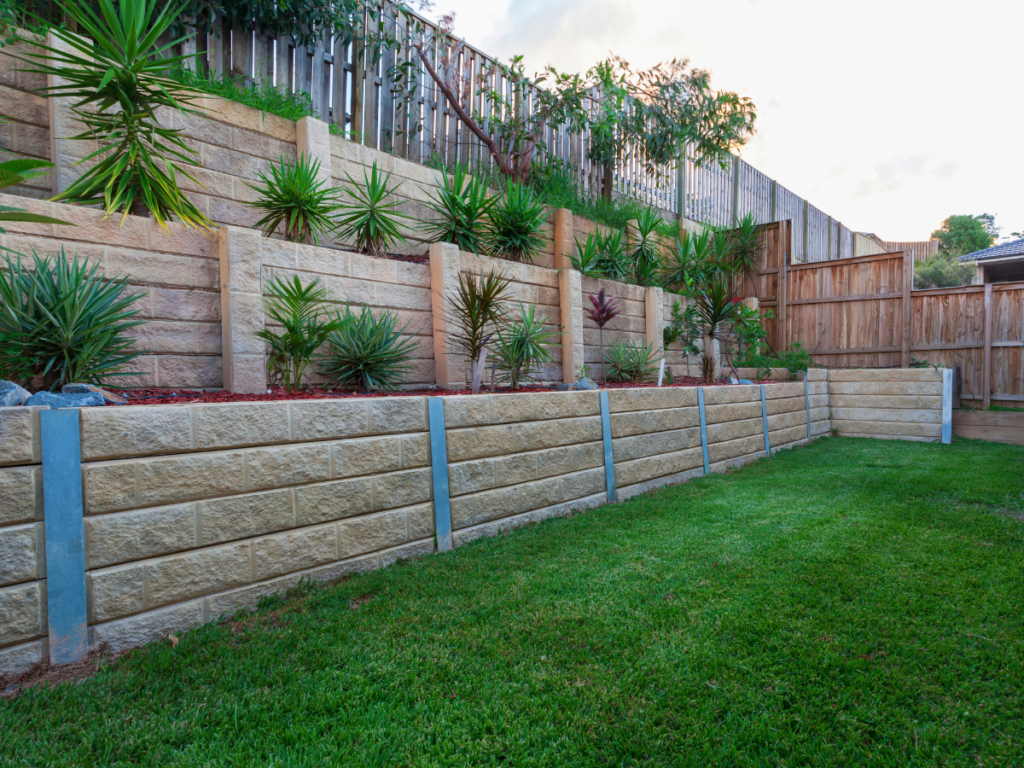 Tiered stone retaining walls with metal supports, landscaped with green plants, border a neatly mowed lawn in a fenced backyard. The robust stone retaining wall not only adds structural strength but also enhances the overall elegance of the outdoor space.
