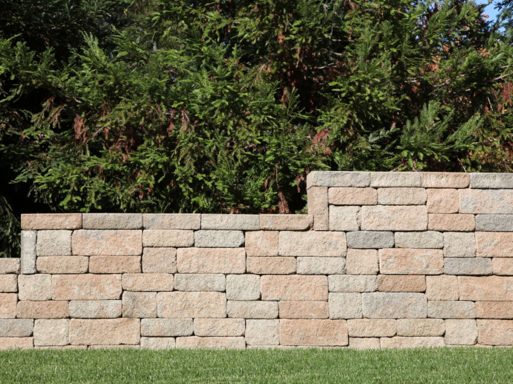 A tan brick and stone retaining wall stands in front of green evergreen shrubs, with neatly trimmed grass in the foreground.