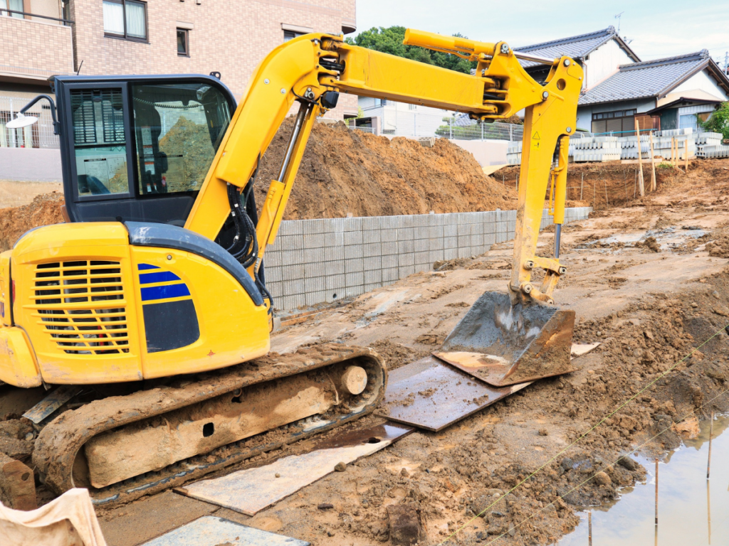 A yellow excavator is parked on a muddy construction site near a partially built stone retaining wall, with houses visible in the background.