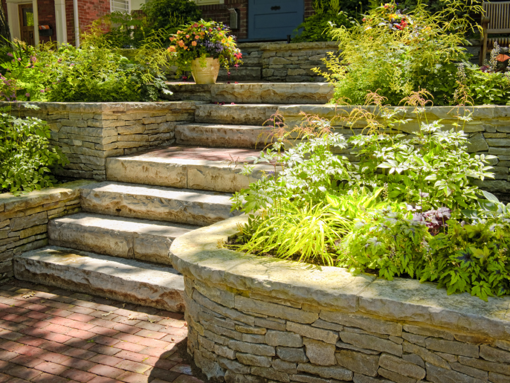 Stone steps bordered by a stone retaining wall, raised garden beds, and lush plants lead up to a brick patio and a porch adorned with potted flowers.