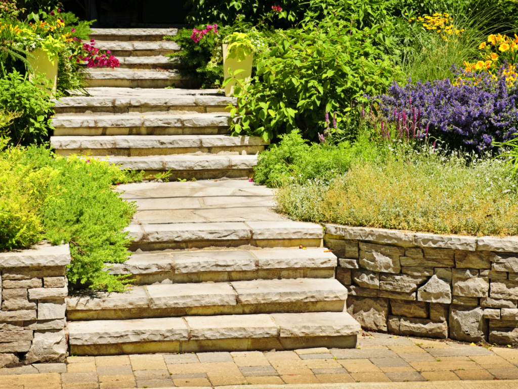 Stone garden steps bordered by green shrubs and colorful flowers lead upward, with a beautifully crafted stone retaining wall and a paver path in the foreground under bright sunlight.