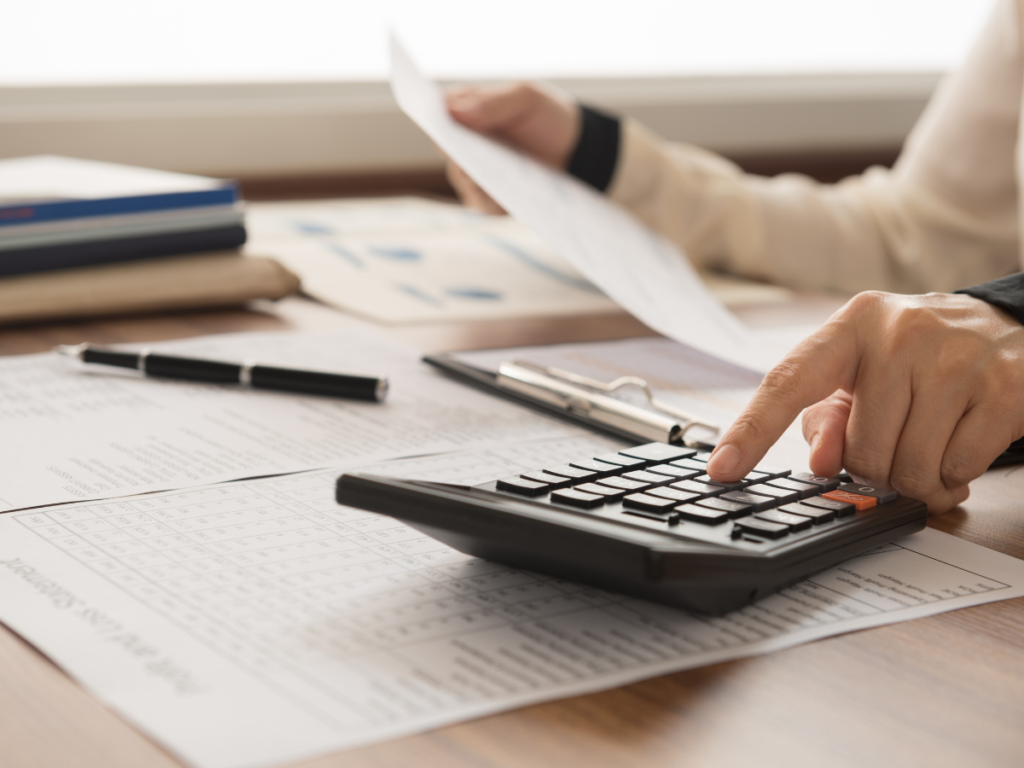 Person using a calculator and holding documents at a desk with papers, a pen, and a clipboard, engaged in financial or accounting work while seated near a window overlooking a stone retaining wall.