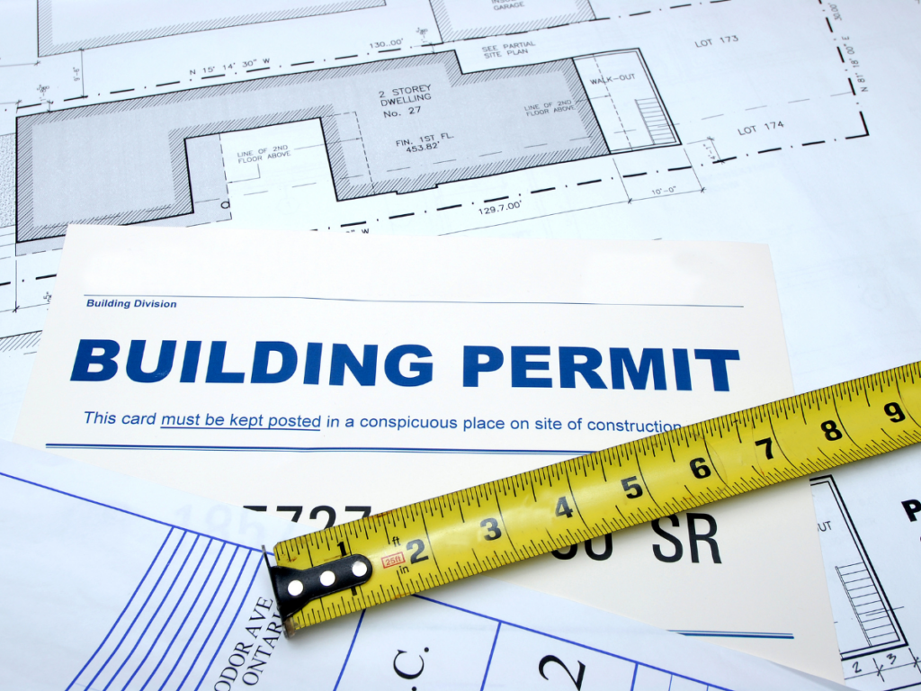 A building permit, measuring tape, and architectural blueprints are shown together, indicating construction planning or approval, possibly for a project such as a stone retaining wall.