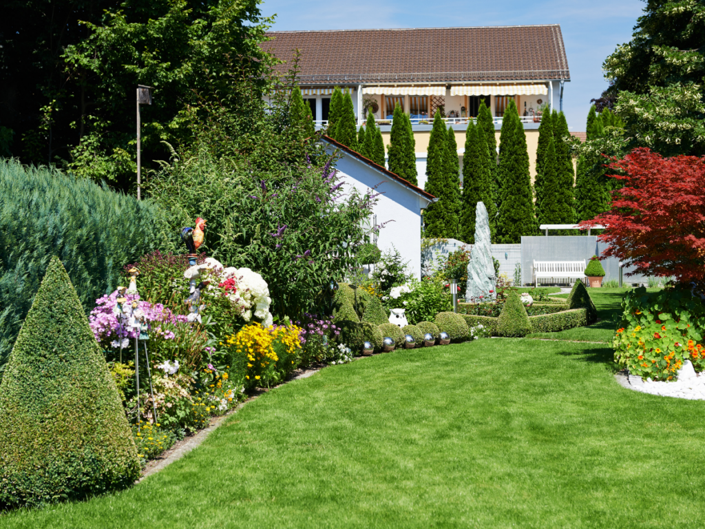 Well-manicured garden with trimmed shrubs, colorful flowers, a lawn, and a house with tall trees in the background on a sunny day—beautifully showcasing 3D landscape designs that bring outdoor spaces to life.